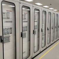 View of an industrial control room featuring rows of electrical panels in a well-lit environment.