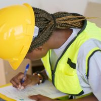 A female engineer in PPE writing on a clipboard at a construction site.