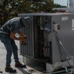 A worker in a bucket hat repairs an outdoor air conditioning unit on a rooftop.