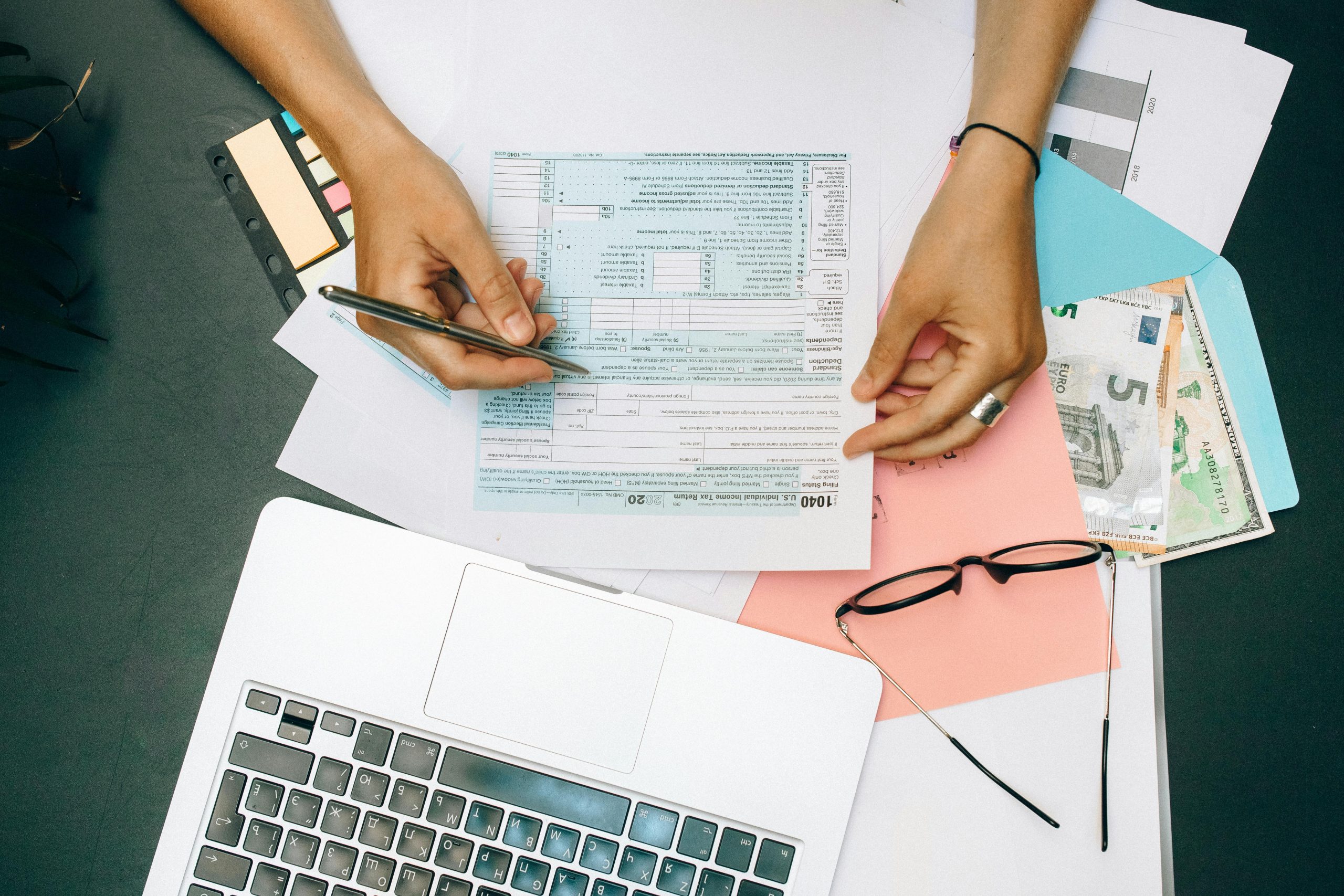 Design, Engineering & Project Management Hands writing on tax documents with laptop, glasses, and currency on desk.